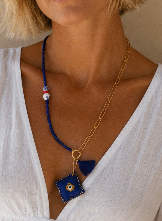 A close-up of a model wearing a multi-material necklace featuring a blue beaded section with white and blue evil eye beads, a gold chain, and a blue fabric square pendant with a gold eye design.